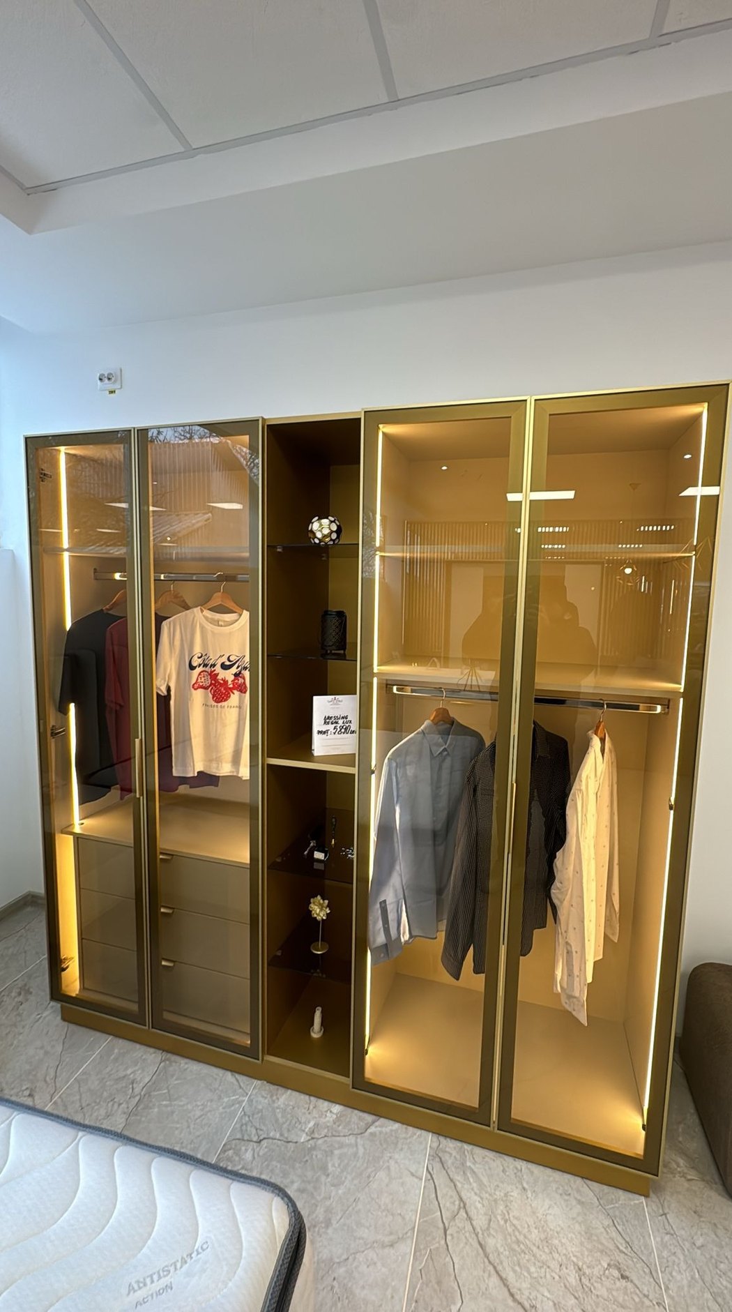 Dulap cu usi de sticla si rama aurie fotografiat cu telefonul in showroom, cu tenta galbena si o saltea in colt